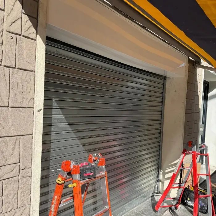 Muntatge d’una persiana metàl·lica al portal d’un comerç, amb dues escales i el calaix superior obert